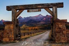 an old wooden gate in the middle of a dirt road with mountains in the background