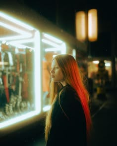 a woman with long red hair standing in front of a store window looking into the distance