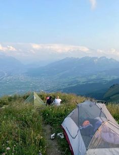 two tents set up on top of a hill with mountains in the background and people sitting outside