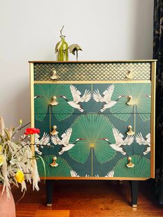 a green and gold dresser with birds painted on the front, flowers in vases next to it