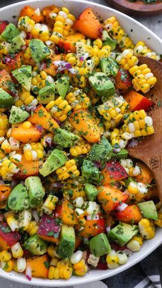 a bowl filled with corn, avocado, and other veggies next to a wooden spoon