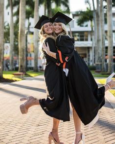 two women in graduation gowns are hugging each other on the sidewalk with palm trees behind them