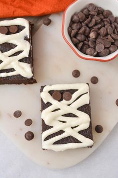 two brownies with white frosting and chocolate chips next to a bowl of cookies