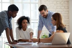 three people standing around a table looking at something on the computer screen while another person looks down