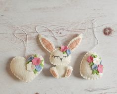 three felt bunny ornaments hanging from strings on a white wooden surface, one with pink and blue flowers in it's ears