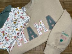 "Mama Embroidered Sweatshirt with baby onsies and name on sleeve | Baby Outfit Keepsake Applique Sweatshirt | Simple Mama Pullover | Gift for Mom, | Personalized Mama Shirt | Mama Gift | Mom Gift | Mama Shirt | Mama Sweatshirt Are those baby outfits piling up because you don't know what to do with them?  The Keepsake Mama Sweatshirt will make the perfect gift for Mama. They're perfect for moms, Mother's Day Gift, or just a simple gesture to say I Love you Mama. We'll use your fabric to create the word \"MAMA\". This design is only for MAMA, if you need another word, please reach out to us prior to placing your order. You also have the option of customizing the sleeve with a name and heart.  IMPORTANT: We CANNOT use parts of the clothing that have vinyl.  Our heating process will remove the Diy Mom Sweatshirt, Childrens Handprint Wreath Sweatshirts, Mama Sweatshirt With Baby Clothes, Mama Embroidery Sweatshirt, Mama Onesie Sweatshirt, Mama Patch Sweatshirt, Custom Mama Sweatshirts, Mama Embroidered Sweatshirt, Momma Embroidered Sweatshirt