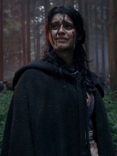 a woman with black hair and makeup standing in the woods wearing a cloak over her shoulders