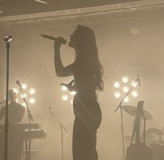 a woman singing into a microphone while standing in front of a band on stage with lights behind her