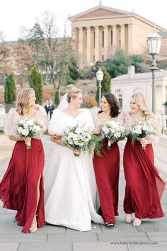 four bridesmaids in red dresses and fur stoles smile at each other while holding bouquets