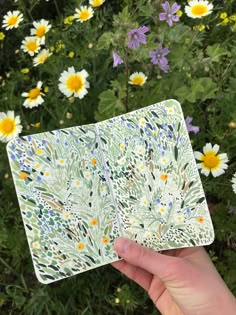 a hand holding up a piece of paper with flowers in the background and daisies