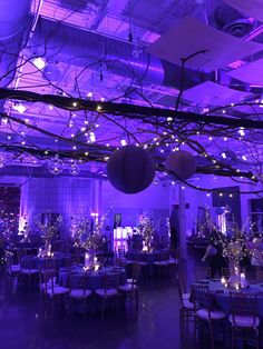 a room filled with lots of tables and chairs covered in purple lights, surrounded by branches