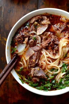 a bowl filled with noodles, meat and vegetables on top of a wooden table next to chopsticks