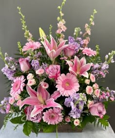 a bouquet of pink and purple flowers in a square vase on a table with greenery