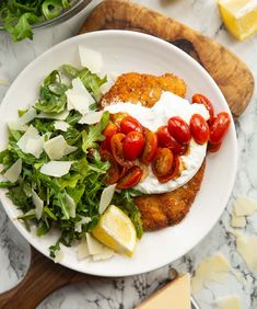 a white plate topped with meat covered in tomatoes and lettuce next to lemon wedges