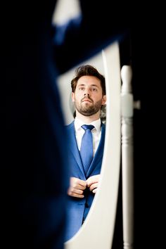 a man wearing a blue suit and tie looking at himself in the mirror with his hands on his hips