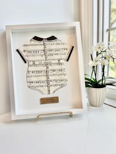 a white frame with sheet music in the shape of a baby's shirt and bow tie