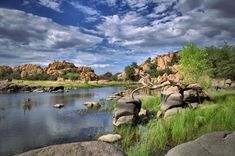 an elephant is standing in the water near some rocks and grass with clouds above it