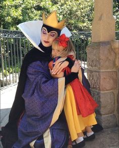 a woman dressed as evil queen holding a doll in front of a fence with trees behind her