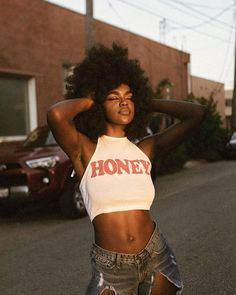 a woman with an afro standing on the street in front of a car and holding her hair back