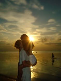 a man and woman hug on the beach as the sun sets in the distance behind them