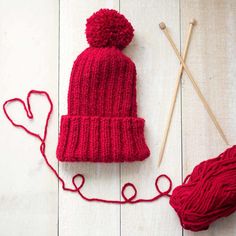 a red knitted hat and two knitting needles on a white wooden surface with yarn