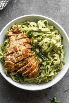 a white bowl filled with pasta and chicken on top of a gray table next to a fork