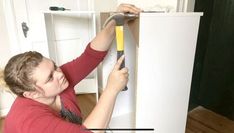 a woman is holding a hammer in front of a white cabinet with the door open