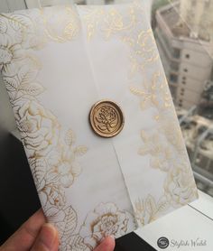 a person holding up a white and gold wedding card with a wax stamp on it