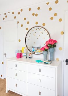 a white dresser topped with flowers next to a wall covered in gold polka dot decals