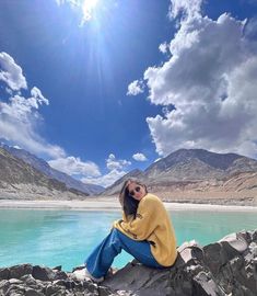 a woman sitting on top of a rock next to a body of water with mountains in the background