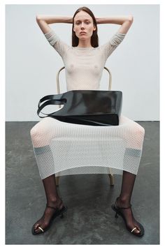 a woman sitting on a chair with her hands behind her head, holding a black bag