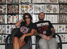 a man and woman sitting next to each other in chairs with tattoos on the wall behind them