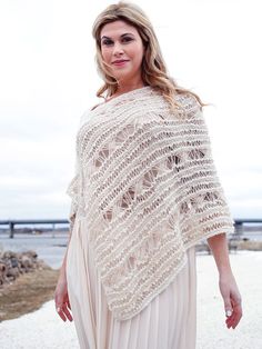 a woman wearing a white crochet shawl standing on the beach with her hands in her pockets