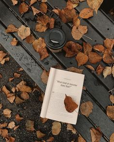 an open book sitting on top of a wooden bench next to leaves and a coffee cup