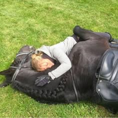 a woman laying on top of a black horse in the middle of some green grass