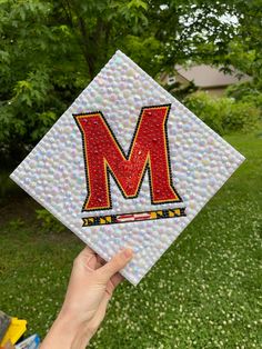 someone is holding up a mosaic tile with the letter m on it in front of some trees