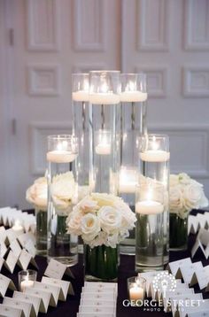 a table topped with lots of vases filled with white flowers and lit votive candles