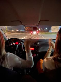 two women driving down the road at night with their hands on the steering wheel and lights on