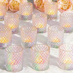 many different colored glass cups sitting on a table with flowers in the backround