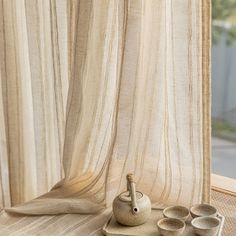 a tea pot and four cups on a table near a window with sheer drapes