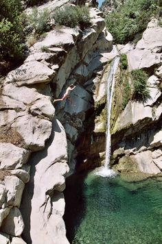 a person jumping off a cliff into the water from a small waterfall that is surrounded by large rocks