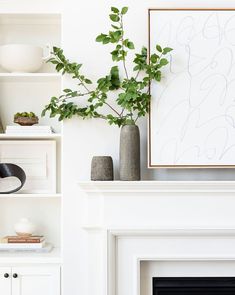 a living room with white furniture and a plant on the fireplace mantel, in front of a framed art piece