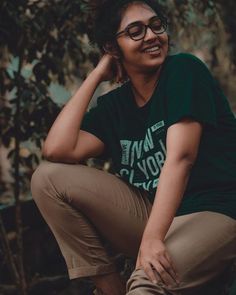 a woman wearing glasses is sitting on a tree branch and posing for the camera with her hands behind her head