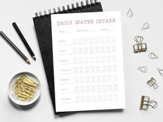 the daily water intake is on top of a notebook next to gold clips and pencils