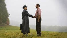 two men shaking hands in the middle of a field with trees and fog behind them