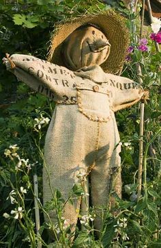 an old scarecrow statue in the middle of some flowers and plants with a straw hat on it's head