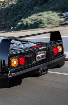 the rear end of a black sports car driving on a road with mountains in the background