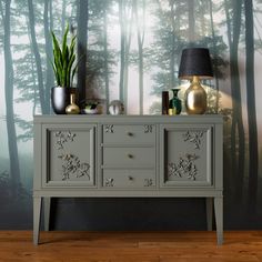 a grey dresser with two vases and a lamp on it in front of a forest wall mural