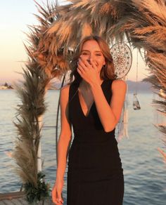 a woman in a black dress standing next to a palm tree and looking at the camera