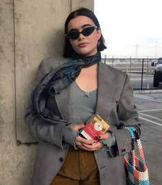 a woman standing next to a wall holding a bag and wearing sunglasses with her hands in her pockets